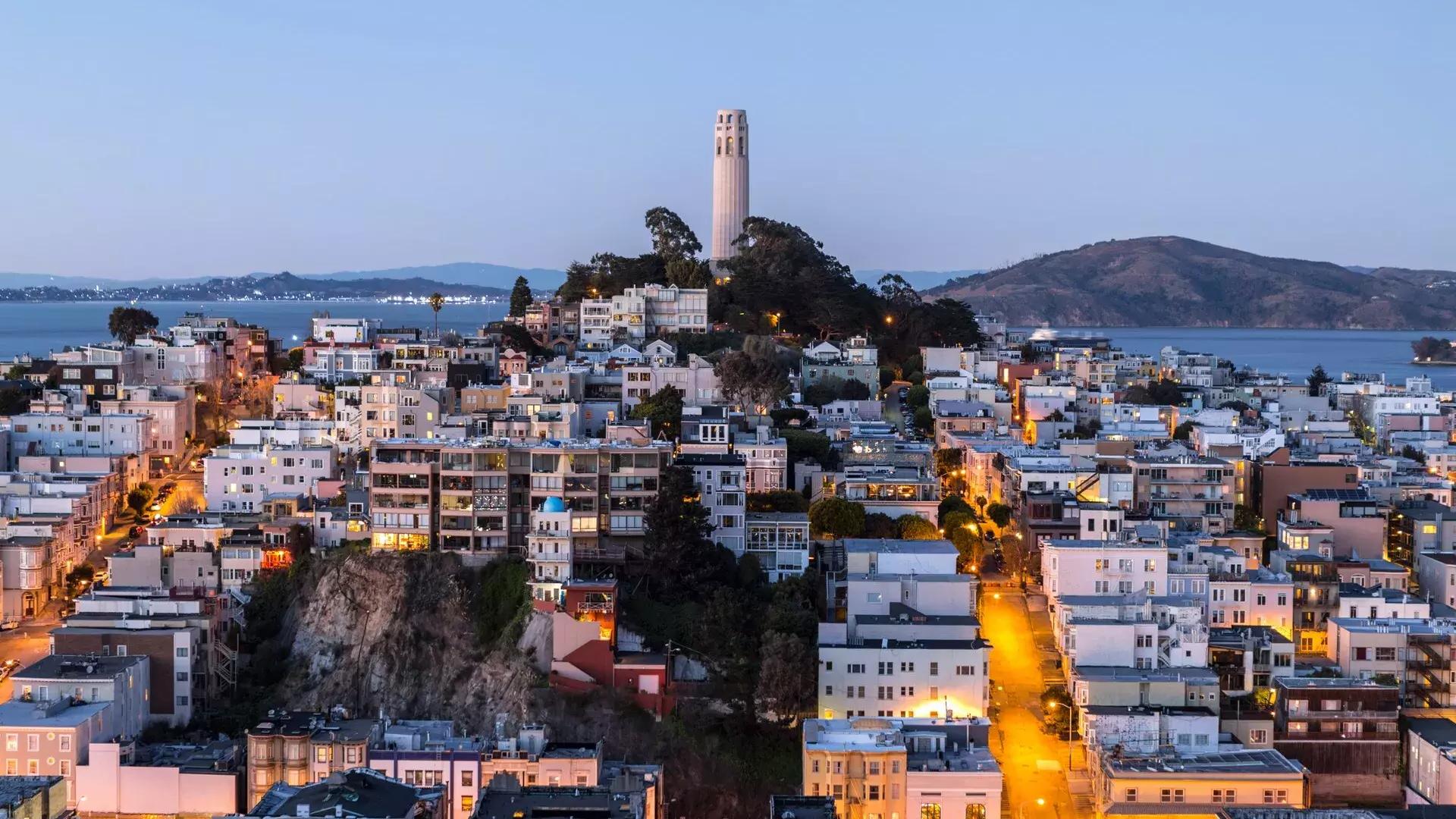 La Torre Coit de San Francisco al anochecer, con calles iluminadas delante y la Bahía de San Francisco detrás.