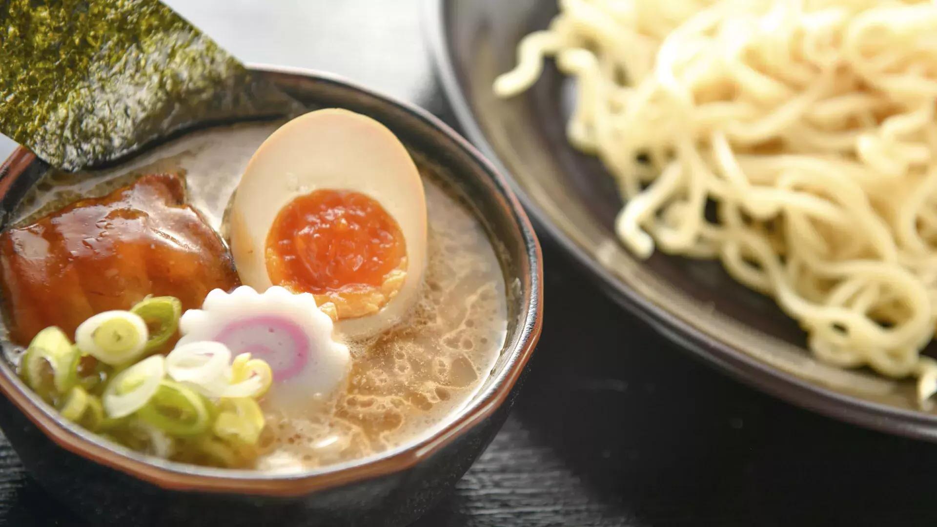 Close up shot of a bowl of noodles and a bowl of ramen soup with a poached egg, sliced in half.