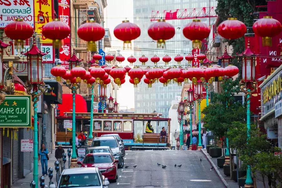 Abgebildet ist eine hügelige Straße in San Franciscos Chinatown mit baumelnden roten Laternen und einer vorbeifahrenden Straßenbahn.