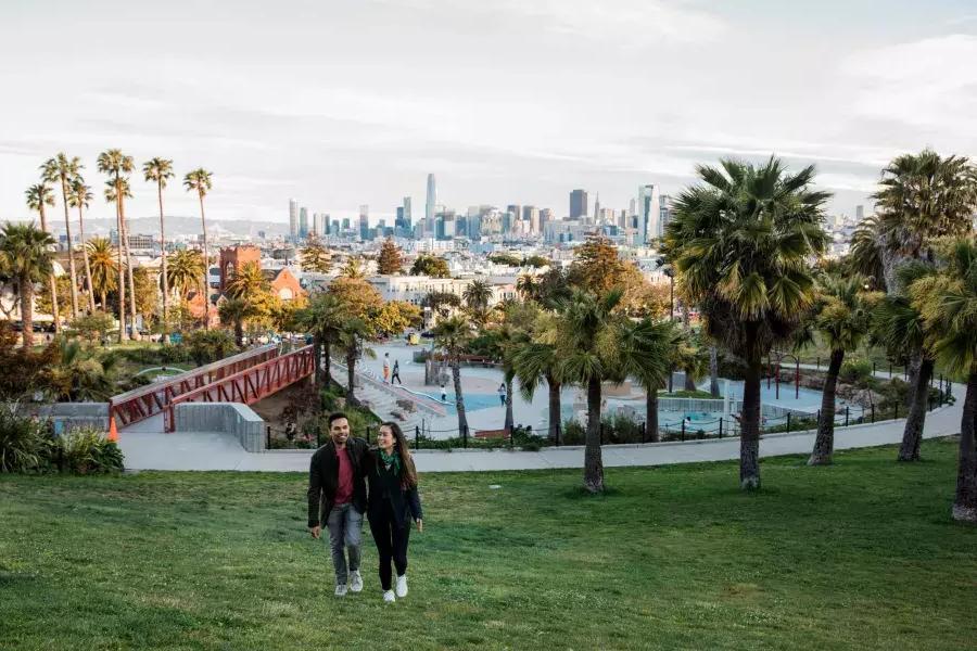 Um casal caminha em direção à câmera com Dolores Park e o horizonte de São Francisco atrás deles.