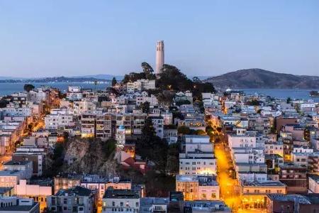 San Francisco's Coit Tower at dusk, with lighted streets before it and the San Francisco Bay behind it.
