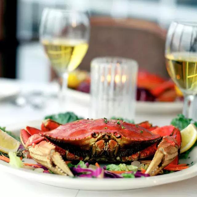 Um caranguejo Dungeness sentado em um prato em um restaurante com duas taças de vinho branco ao fundo.