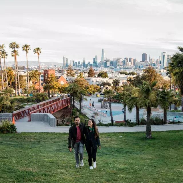 Una pareja camina hacia la cámara con Dolores Park y el Skyline de San Francisco detrás de ellos.